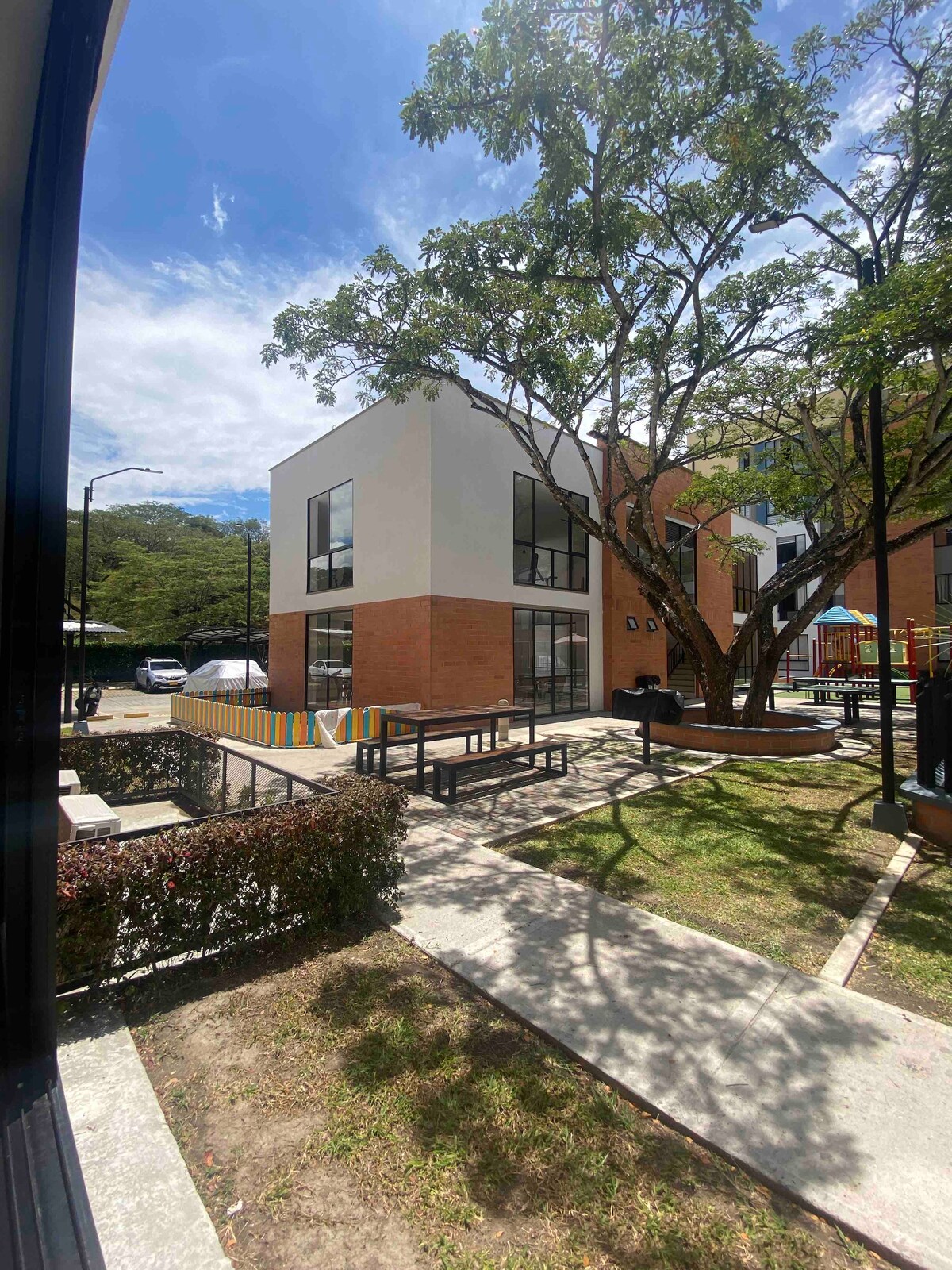 A modern two-story building features large windows that allow for ample natural light. Surrounding green spaces include seating areas and playground equipment, providing options for relaxation and recreation. A tree offers shade in the outdoor environment, enhancing the overall appeal of the residential complex.