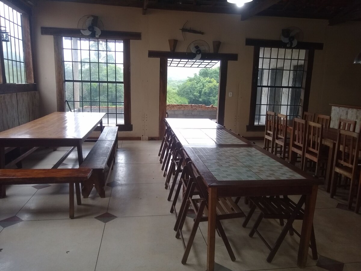 A spacious event area is shown with large wooden tables and chairs arranged for gatherings. Large windows provide natural light and views of the surrounding greenery. Ceiling fans contribute to airflow in the room, enhancing comfort for guests.
