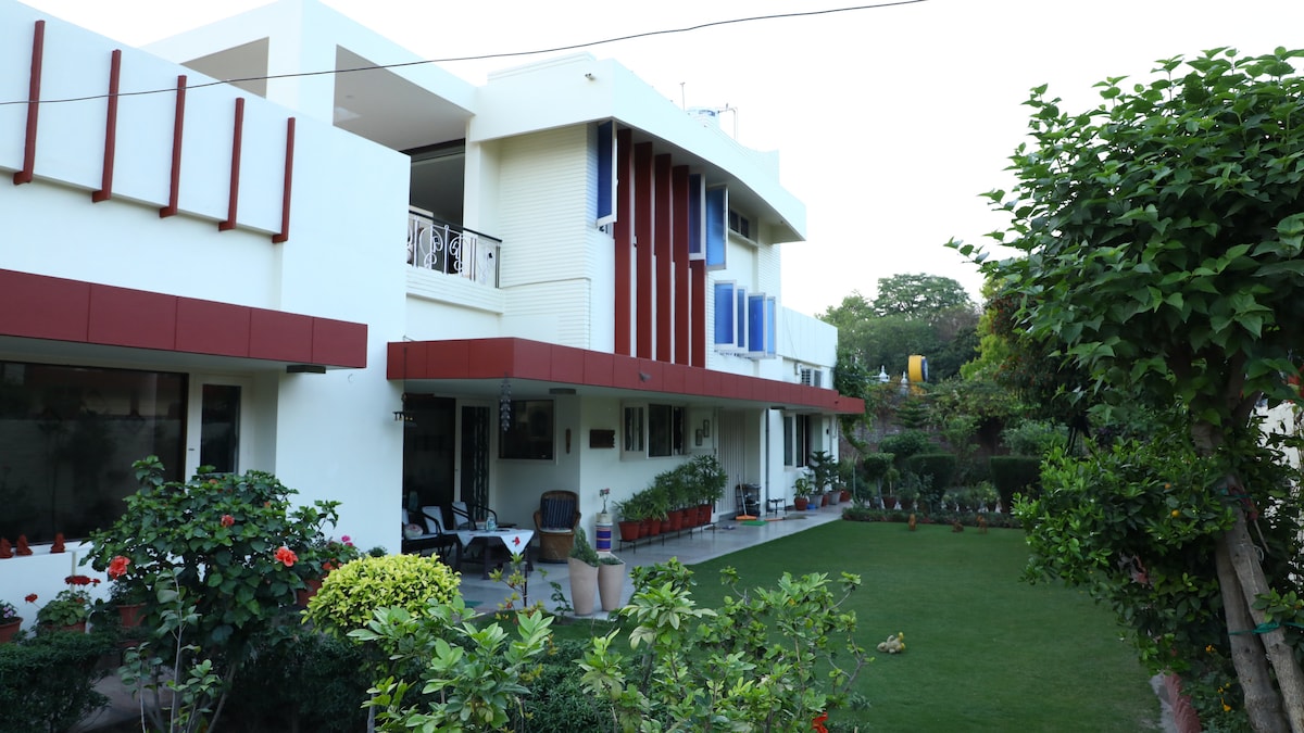 The exterior view of the property highlights a well-maintained garden with lush greenery and colorful flower beds. The building features a modern design with red and blue accents, large windows, and a spacious patio area that invites outdoor relaxation.
