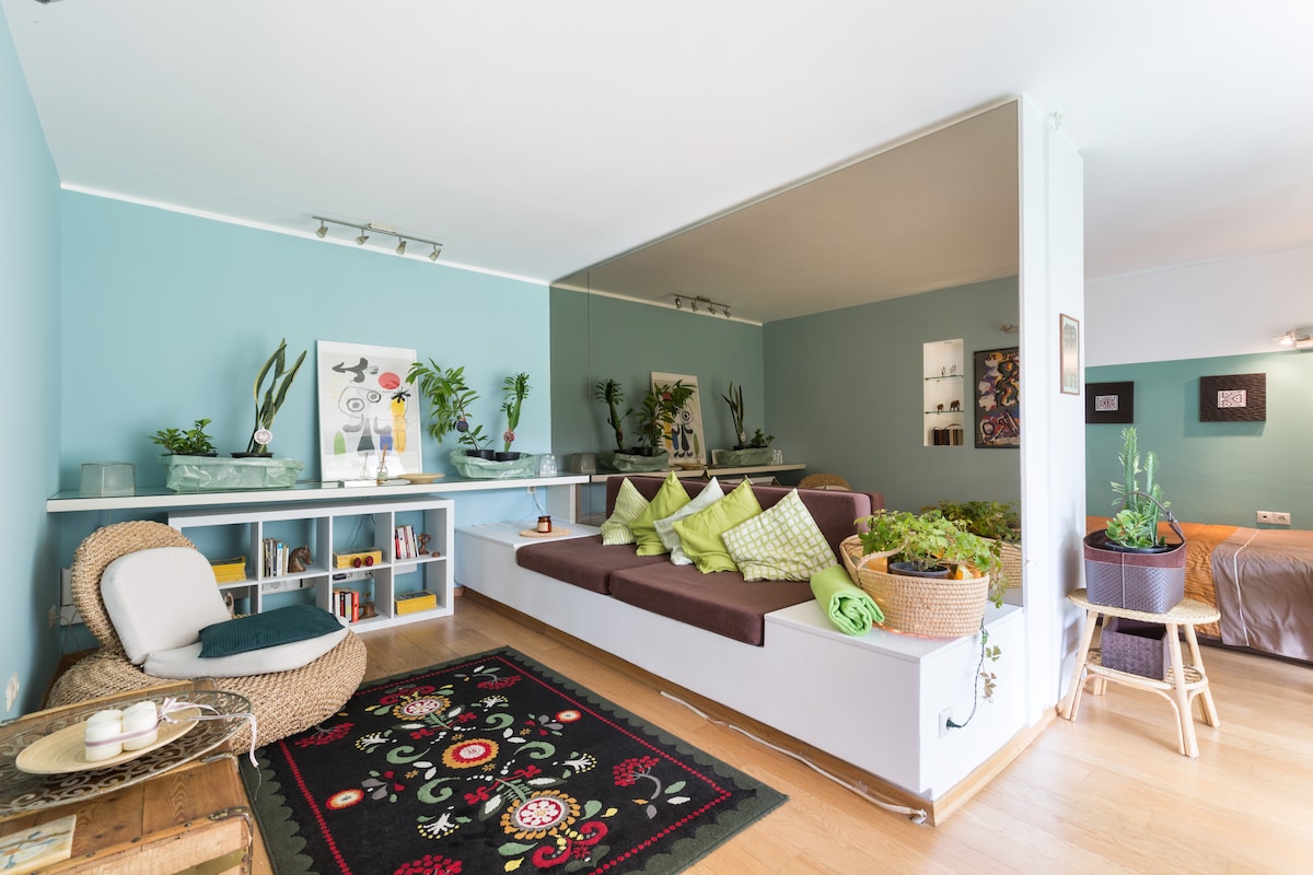 A spacious living area features a large, comfortable seating arrangement adorned with cushions. A vibrant area rug adds color to the light wooden floor. Shelving displays books and decorative items, while plants are positioned throughout, creating a fresh ambiance. A mirrored wall enhances the sense of space.