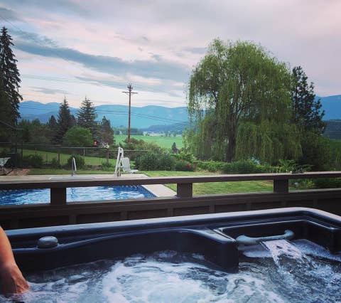 A relaxing hot tub is positioned on a deck, overlooking expansive green fields and distant mountains. Nearby, a swimming pool is visible, surrounded by trees and landscaped gardens, with a cloudy sky creating a serene atmosphere.