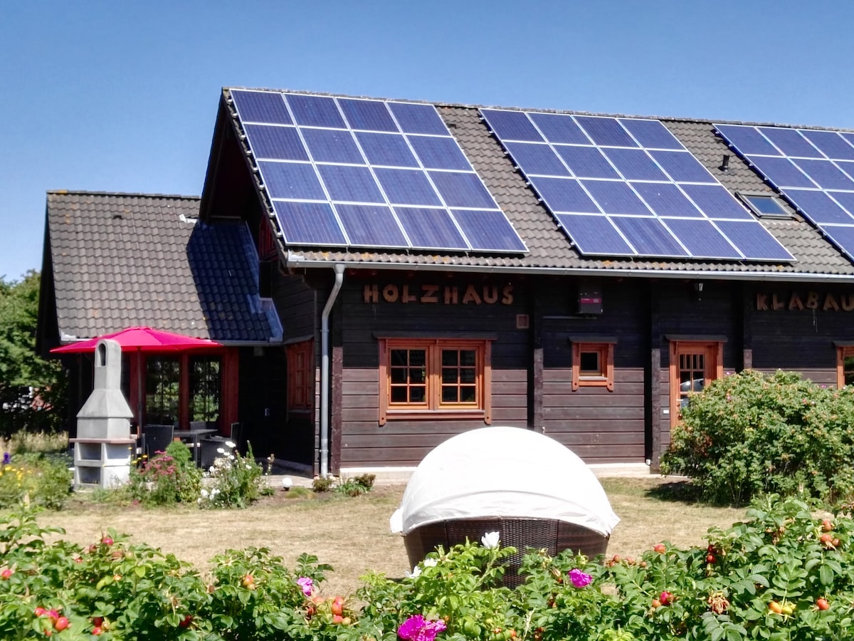 The exterior of the wooden house is showcased, featuring solar panels on the roof. A red parasol provides shade over a sitting area in the garden, which is populated with vibrant flowers. The structure has windows with wooden accents, enhancing its rustic charm.