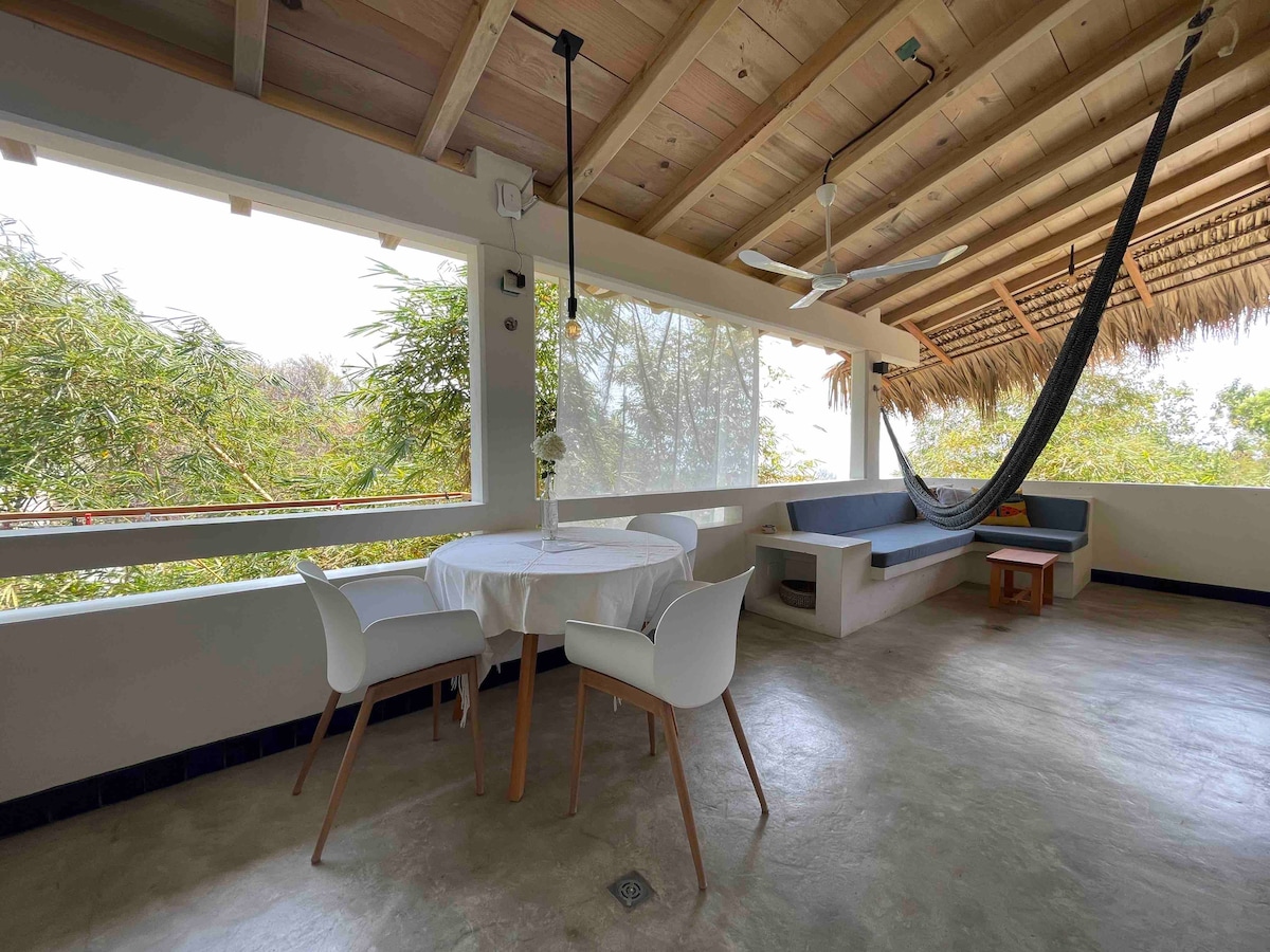 An open-air dining area features a round table with a vase at its center. Surrounding the table are four white chairs. A comfortable seating area with a built-in couch is adjacent, complemented by a large hammock hung across the space. Glass windows provide views of the surrounding greenery.