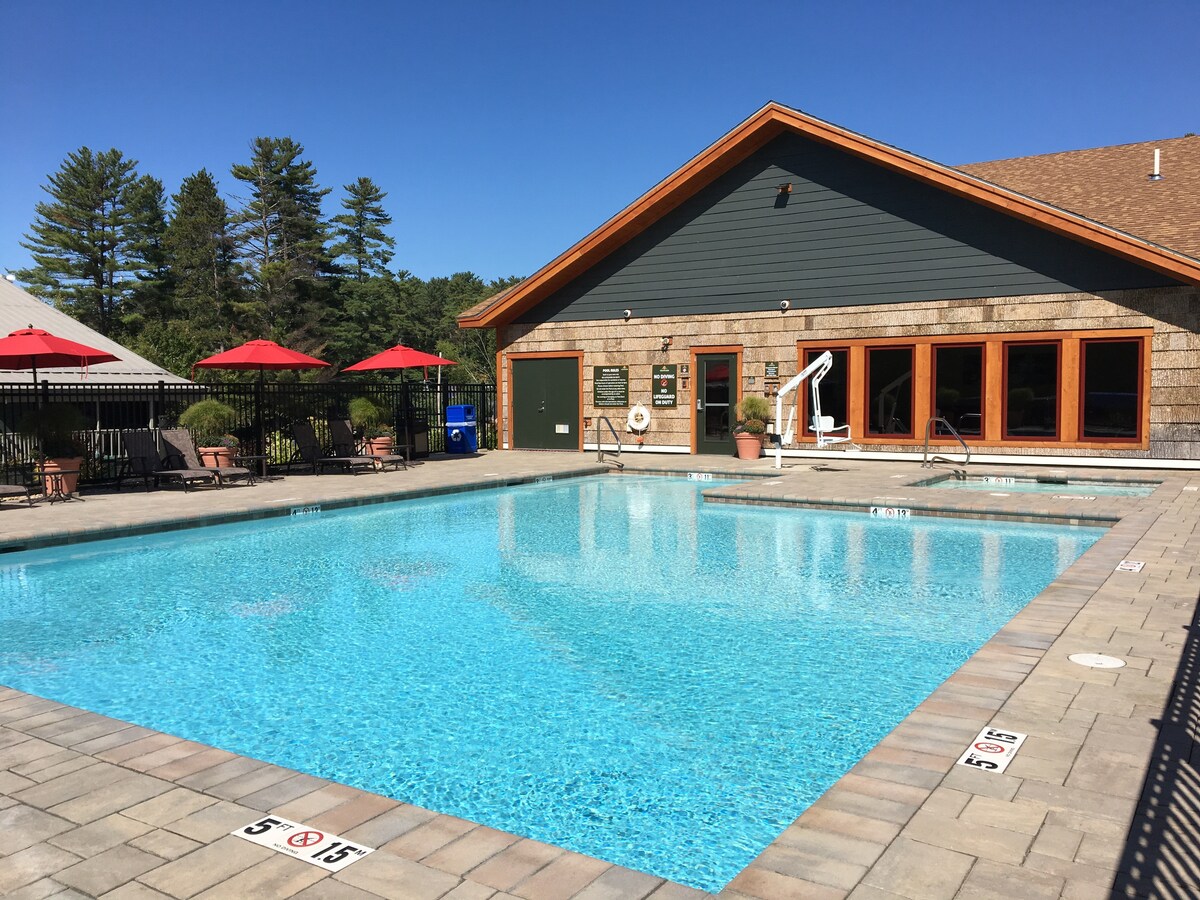 A large outdoor heated pool glistens under a clear blue sky, surrounded by a paved deck with sun loungers and red umbrellas. A building with large windows is situated nearby, offering a welcoming space for relaxation and social gatherings.