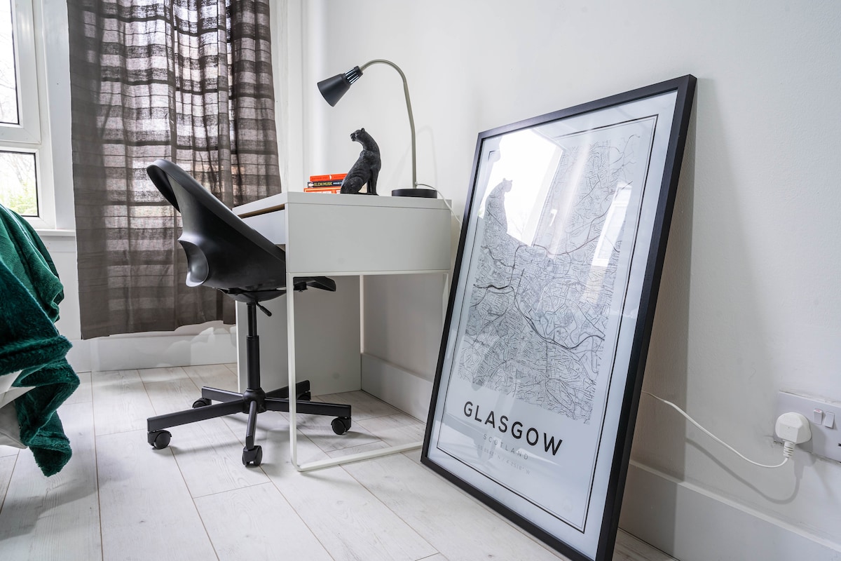 A workspace is presented with a sleek desk and a black office chair. A modern desk lamp illuminates the area. A framed map of Glasgow leans against the wall beside the chair. The room features light-colored flooring and large windows dressed with dark curtains.