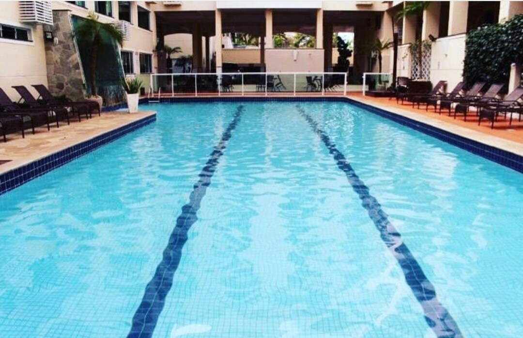 A large swimming pool features clear blue water, with dark blue lane markings visible along the center. Sun loungers are arranged on the pool deck, and the surrounding area is enhanced by tropical plants and architectural elements of the building.