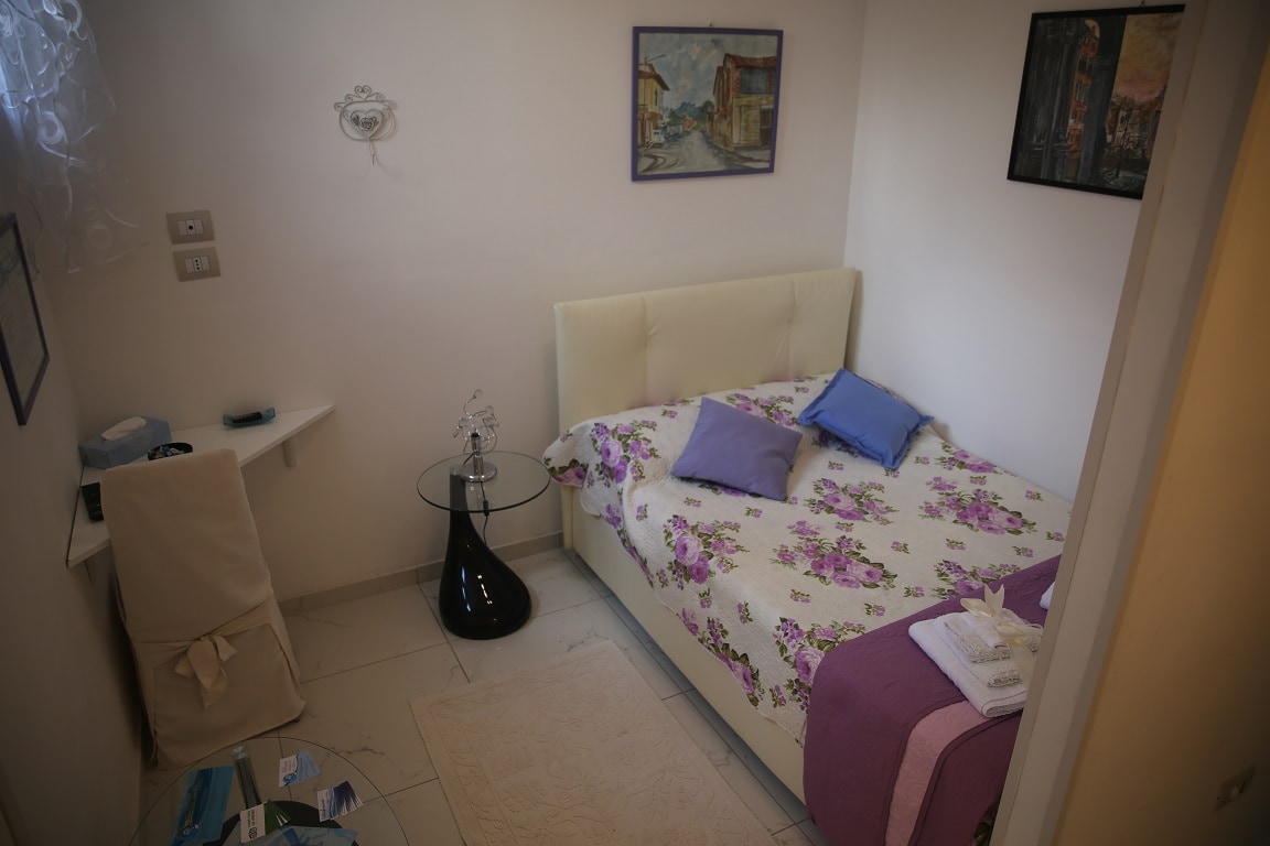 A comfortable sleeping area features a spacious bed adorned with a floral quilt. A small glass table and a chair are positioned nearby. Decorative wall art contributes to the serene atmosphere, while natural light enters through a window draped with sheer curtains.