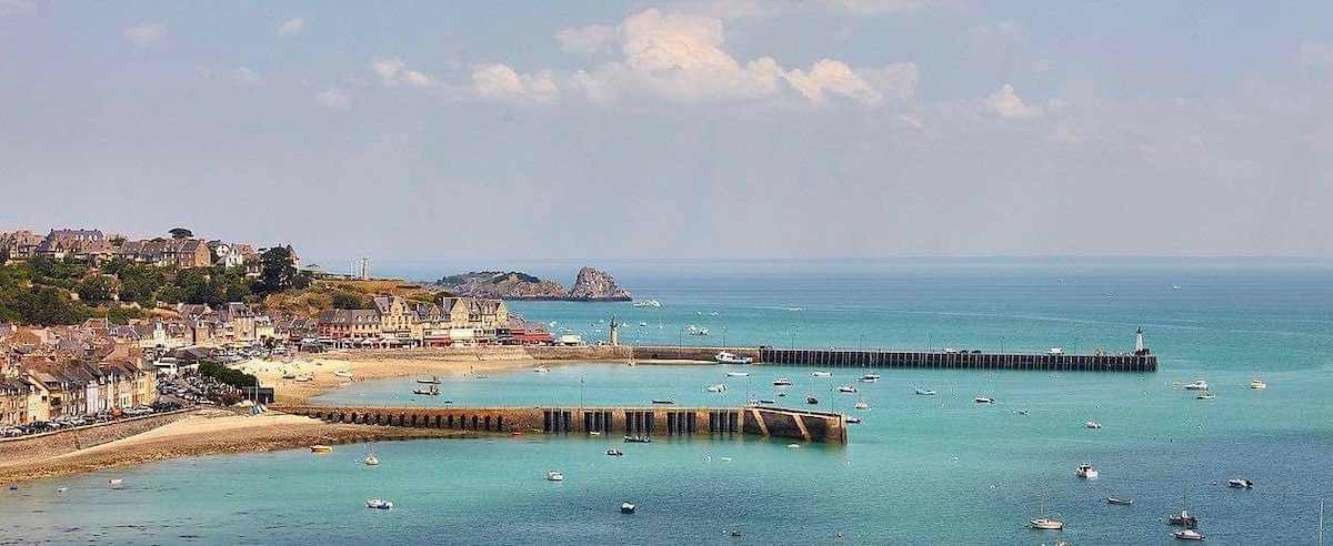 A scenic view of a coastal area features a sandy beach lined with small boats. The calm blue water reflects the sky, and a series of piers extend into the sea, with the shoreline visible in the background. The distant hills provide a natural backdrop.