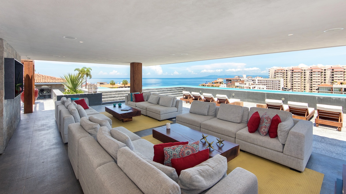 An expansive lounge area features multiple light gray sofas arranged around a wooden coffee table. Bright red accent pillows add color. Large windows provide a panoramic view of the sea and city, with sun loungers situated nearby on a spacious terrace.