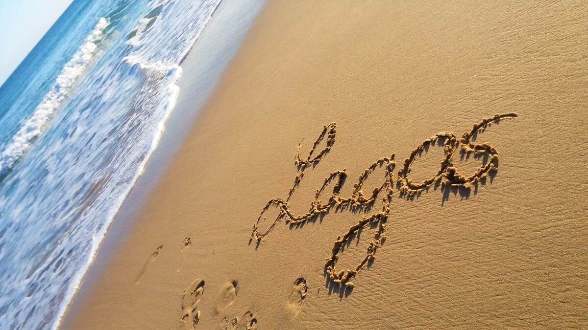 The sandy shoreline features the word 'Lagos' delicately drawn in cursive, with gentle ocean waves softly lapping at the edge. Bright sunlight reflects off the water, creating a serene coastal ambiance.