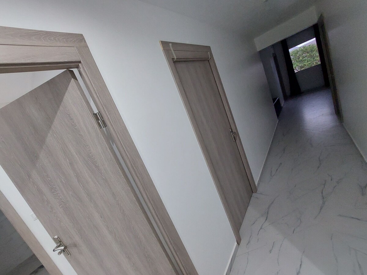 A modern corridor features light-colored walls and a sleek marble floor. Two wooden doors with minimalistic designs are positioned along the hallway. Natural light enters from a window at the far end, illuminating the space.