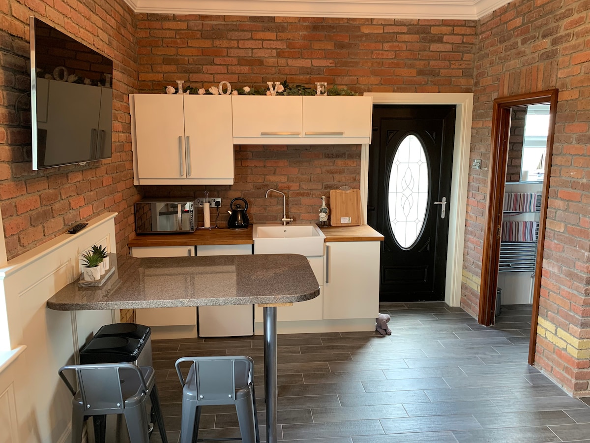 A modern kitchen space features a mix of brick and sleek cabinetry, with a stainless steel sink and a wooden countertop. A flat-screen television is mounted on the wall. Bar stools are positioned at the counter, and a black door provides access to the exterior.