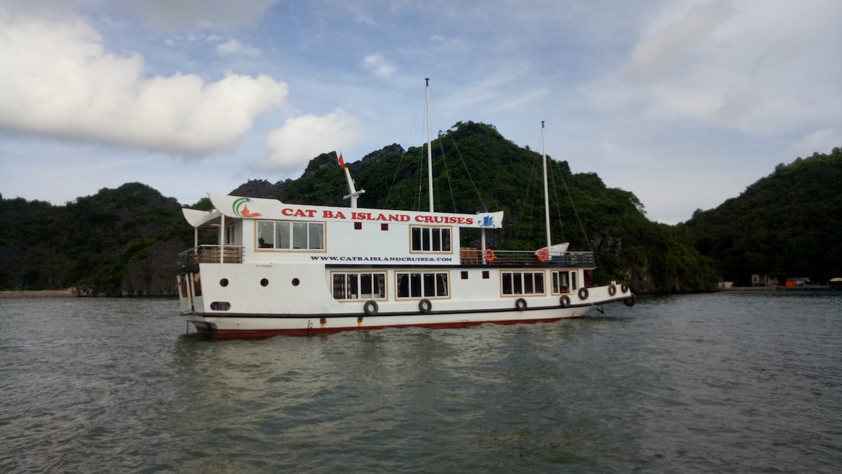 A wooden cruise boat is anchored in the calm waters of Lan Ha Bay, surrounded by lush green hills. The boat features large windows and multiple flags atop its deck, showcasing the name 'Cat Ba Island Cruises' prominently on its side.