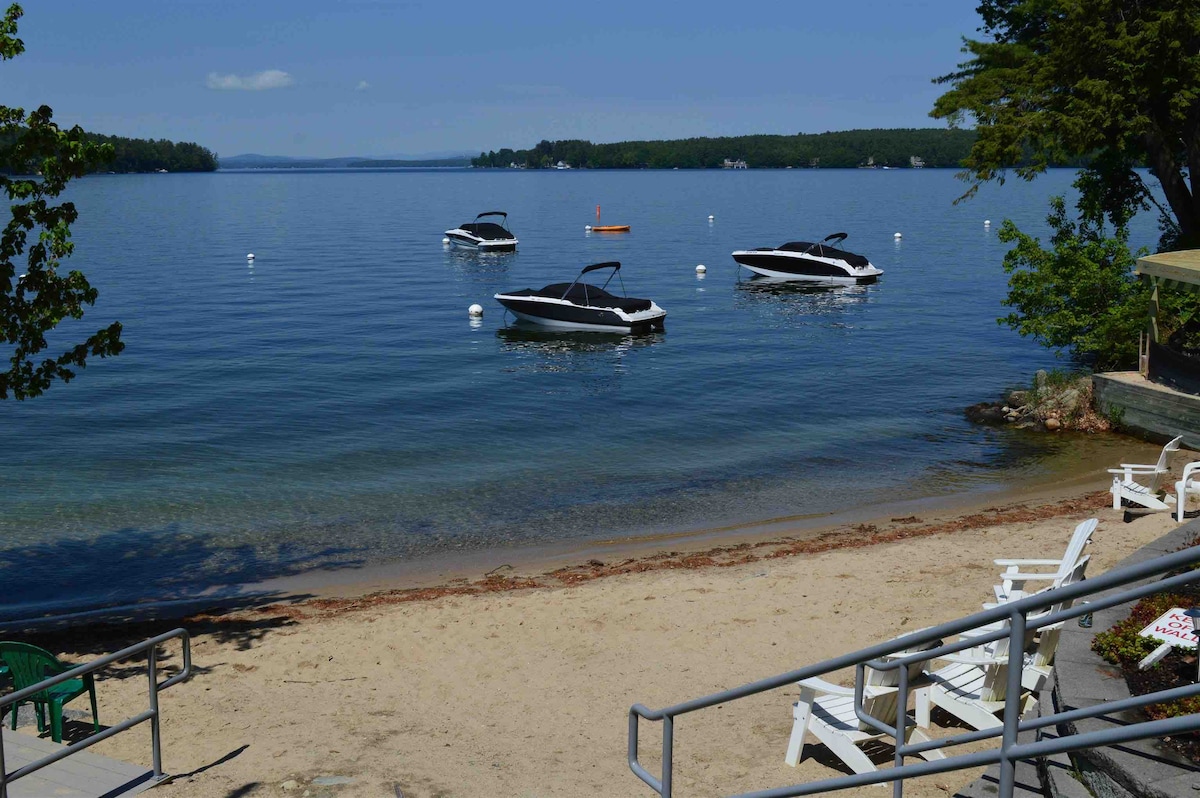 A serene beachfront scene is depicted, featuring several boats anchored on calm waters. A sandy shore extends into the lake, with a few wooden lounge chairs positioned nearby. Lush trees provide shade along the water's edge, creating a peaceful setting for relaxation.