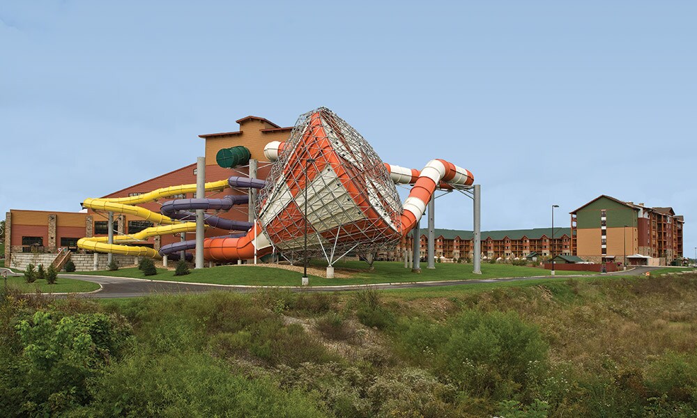 A large water park structure is prominently displayed, featuring colorful water slides in various shapes that extend from a central building. The setting includes lush greenery and open space, with the lodge visible in the background against a clear sky.