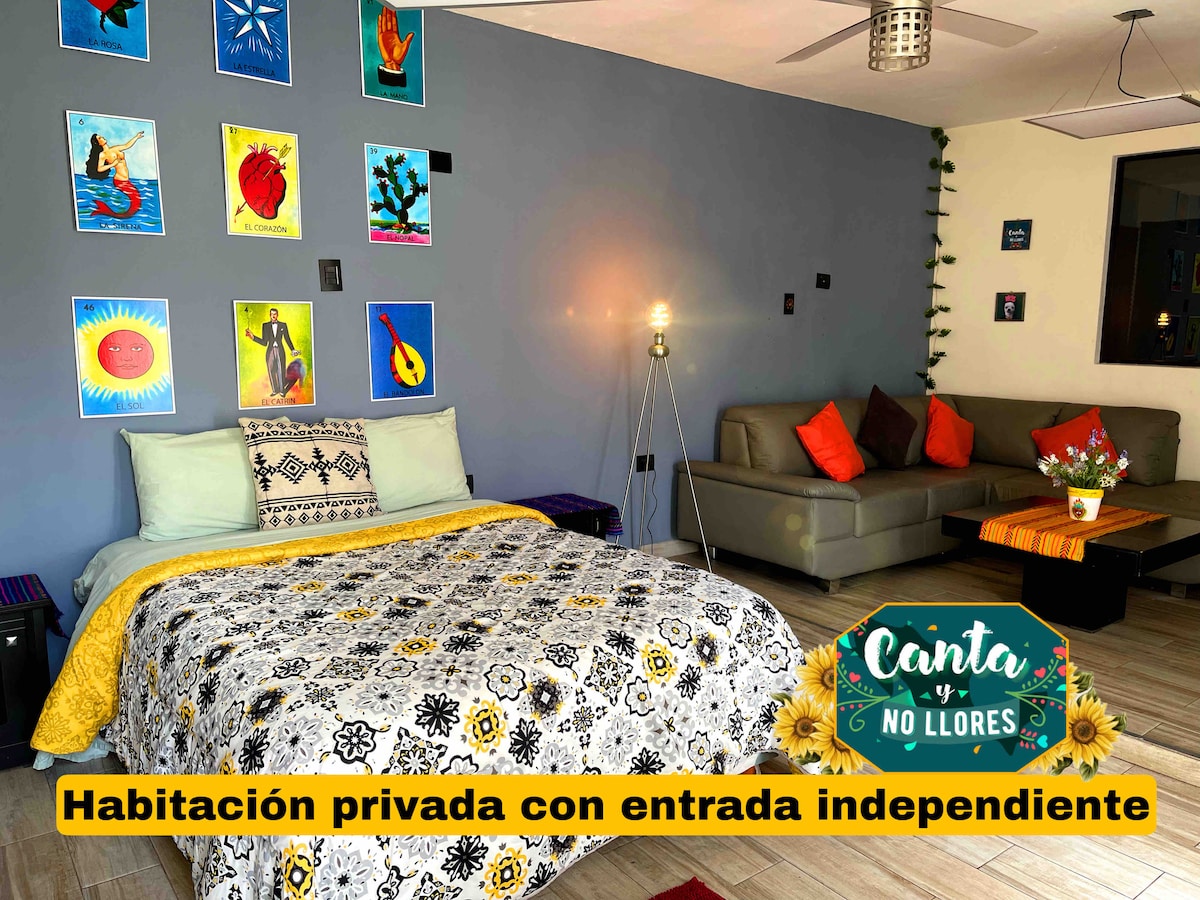 A colorful private room features a spacious bed adorned with a patterned blanket. A comfortable seating area is situated nearby, complemented by wall art depicting musical themes. Natural light enters from the window, creating a vibrant yet cozy atmosphere.