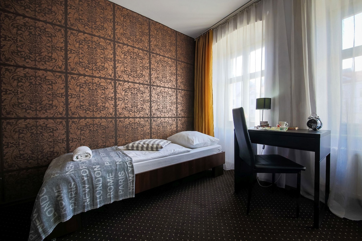 A twin room features two twin beds positioned against an ornate wall with a textured design. A small desk and chair are arranged beside a window dressed in sheer curtains, allowing natural light to fill the space. A light blanket and rolled towel provide a neat touch.
