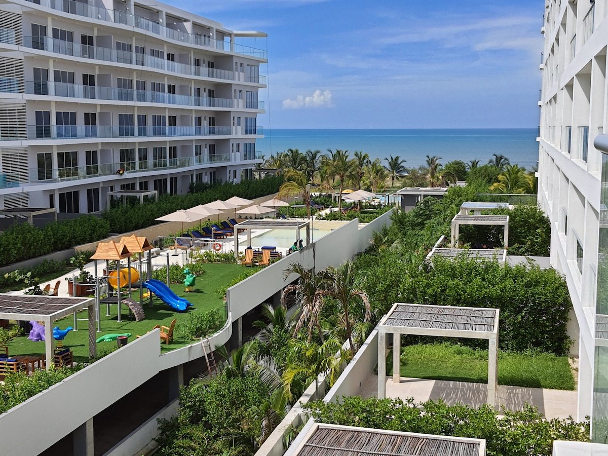 A view of the outdoor recreational area features lush green landscaping, playground equipment, and multiple seating areas. Umbrellas provide shade over lounging spaces. The turquoise sea can be seen in the background, enhancing the tranquil atmosphere of the surroundings.
