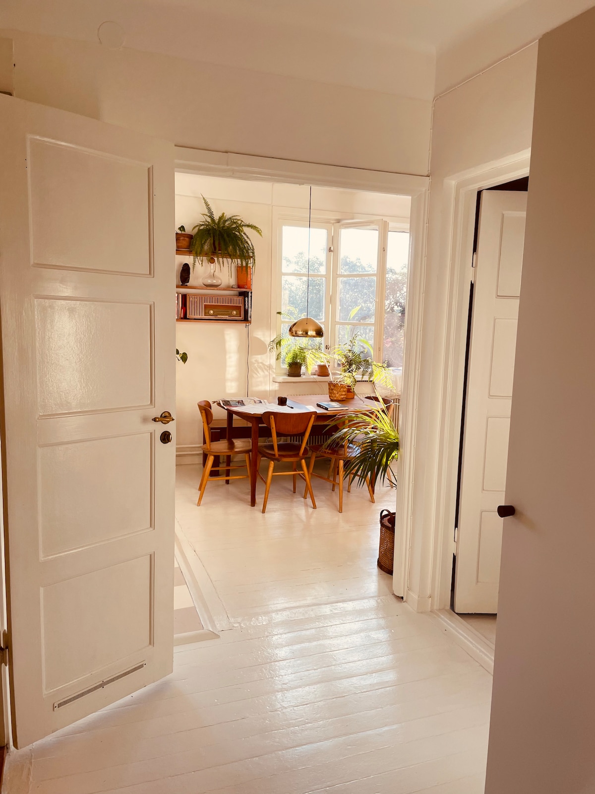 An inviting corridor leads to a brightly illuminated dining area, featuring a wooden table surrounded by four chairs. Potted plants add greenery to the space, and sunlight filters through large windows, enhancing the warm atmosphere of the room.