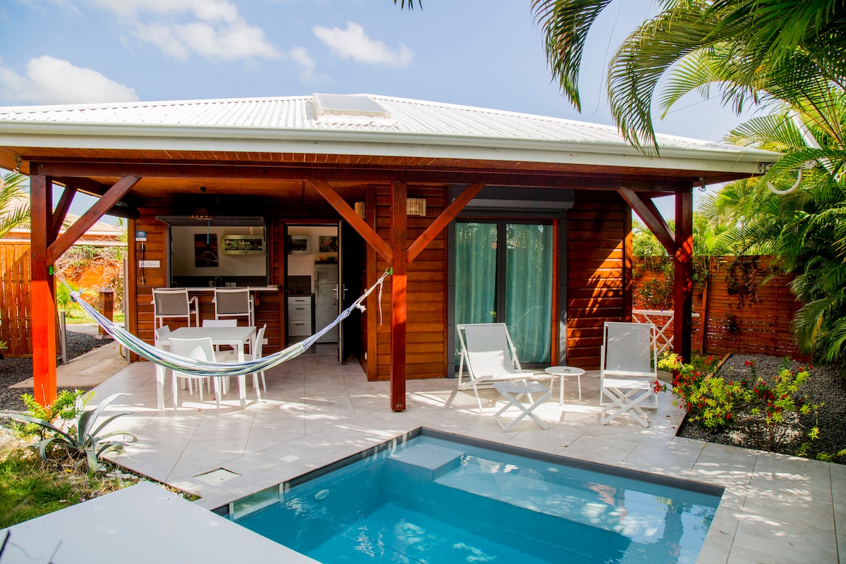 The exterior of the villa displays a covered terrace with a light-colored tile floor and wooden beams. Two white lounge chairs and a hammock are arranged near a private plunge pool, surrounded by tropical plants and greenery. The entrance into a cozy living area is visible behind.