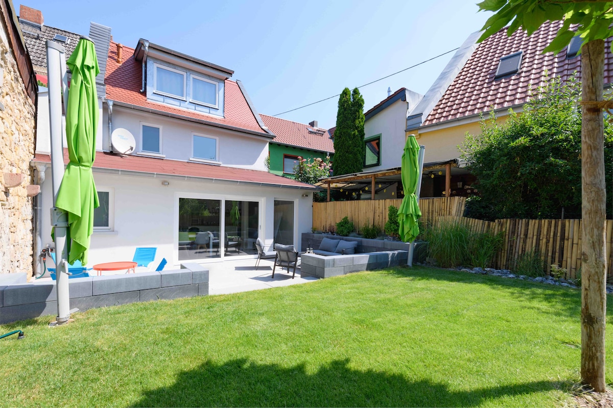 The image depicts a well-maintained garden area adjacent to a charming house. A spacious lawn is complemented by modern outdoor furniture in a seating area. Bright green umbrellas provide shade, and a stone seating area enhances the ambiance. Lush greenery frames the space, creating a peaceful outdoor environment.