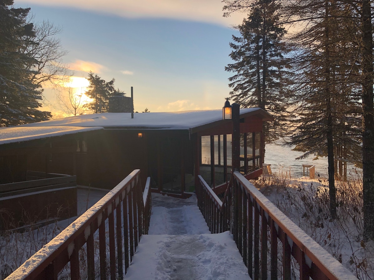A wooden walkway leads to a cabin, surrounded by snow-covered trees, with the sun setting in the background. The cabin features large windows, and a warm light is visible from the entryway, indicating a cozy interior atmosphere.