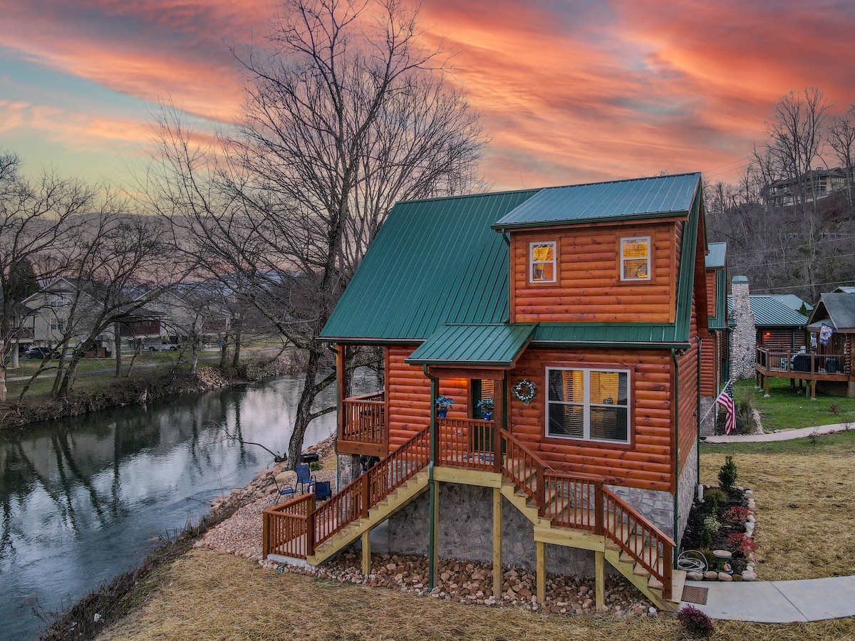 The exterior of a rustic cabin is shown, featuring a green metal roof and wooden siding. It overlooks a calm river, with trees lining the banks. A pathway with steps leads to the front entrance, surrounded by a neatly maintained yard.