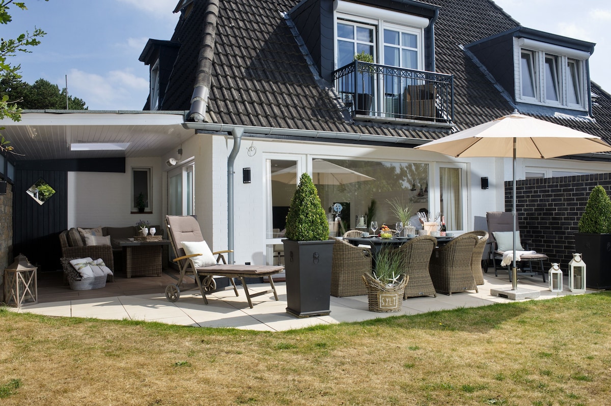 The outdoor terrace features comfortable lounge furniture arranged under a large umbrella, offering shade and relaxation. A dining table is set for meals, with decorative plants and lanterns enhancing the space. The exterior of the house showcases a traditional design, complemented by a small balcony.