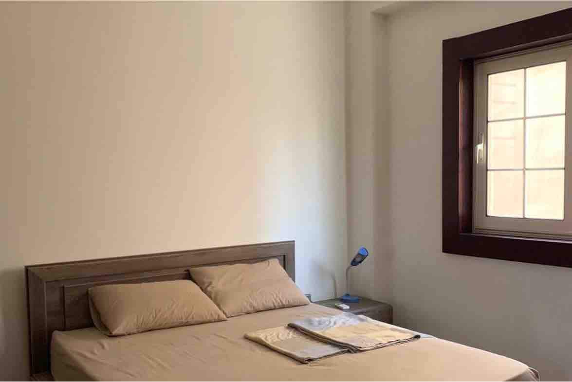 A bright bedroom features a spacious bed with soft-colored linens. A nightstand is positioned beside the bed, adorned with a small lamp. Natural light enters through a square window, enhancing the simple and serene atmosphere.