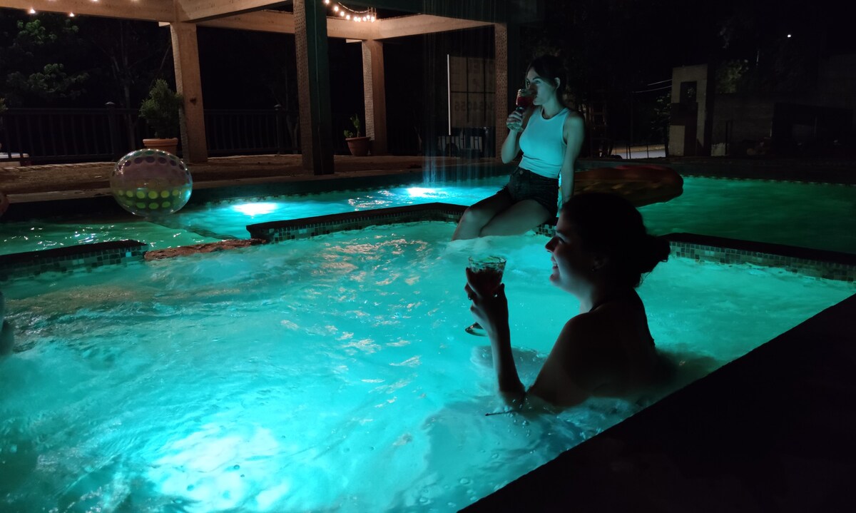 A relaxing evening scene is captured at a pool surrounded by illuminated surfaces in turquoise hues. The water is gently rippling, reflecting the ambient lighting, while a vibrant beach ball adds a playful touch. Seating areas are visible, enhancing the inviting atmosphere for guests.