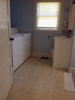 A laundry room features a washing machine and dryer side by side against a light blue wall. A white cabinet provides additional storage space, while a small wastebasket is placed nearby. Natural light filters through a window with horizontal blinds.