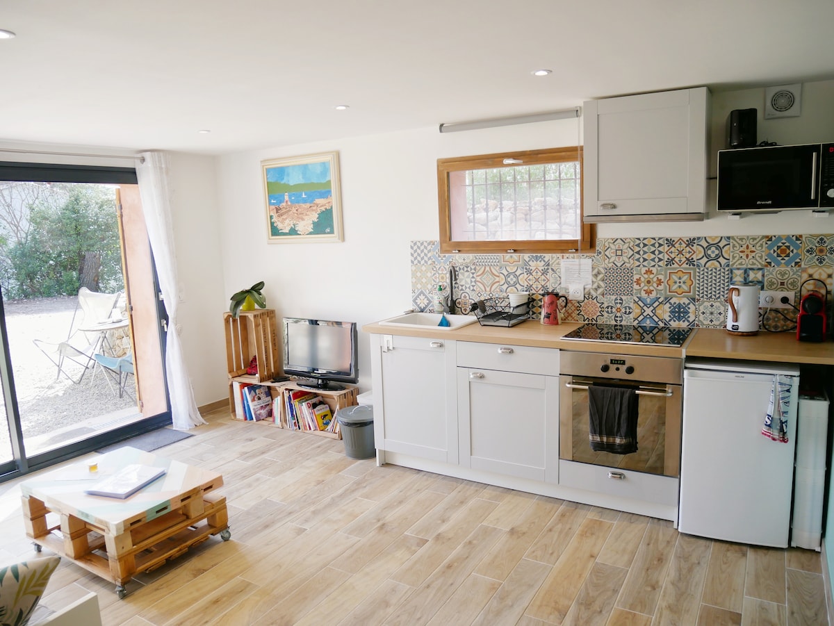 A bright kitchen area features modern appliances, including an oven, stovetop, and refrigerator, set against colorful tiled backsplash. Natural light enters through sliding glass doors, which lead to an outdoor seating area. Wooden shelves display various books, enhancing the cozy atmosphere.