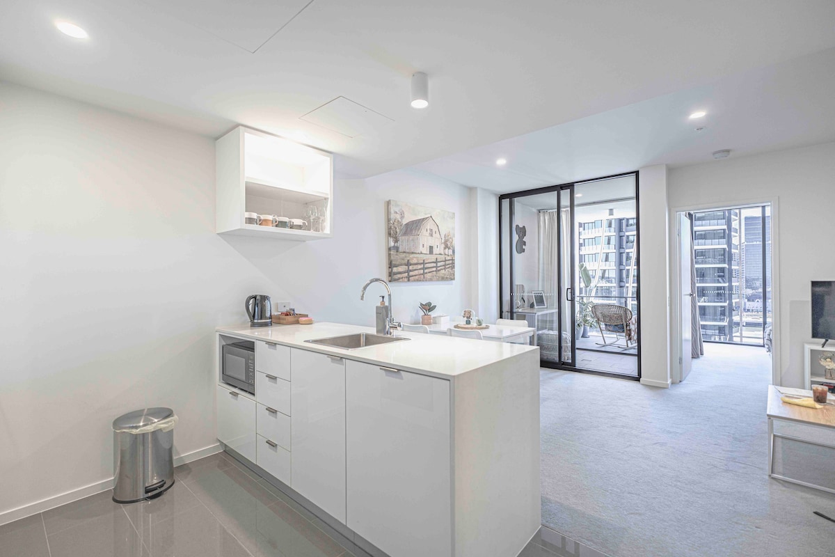 A modern kitchen with sleek white cabinetry and a polished countertop is shown. A microwave and kettle are visible, while a spacious living area with large glass doors opens to a balcony. Natural light brightens the room, enhancing the contemporary design.