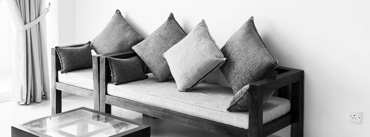 A modern seating area is depicted, featuring a comfortable wooden sofa adorned with gray cushions. Natural light is allowed to filter through a nearby window, highlighting the minimalist design. A glass coffee table sits in front of the seating, complementing the overall aesthetic.