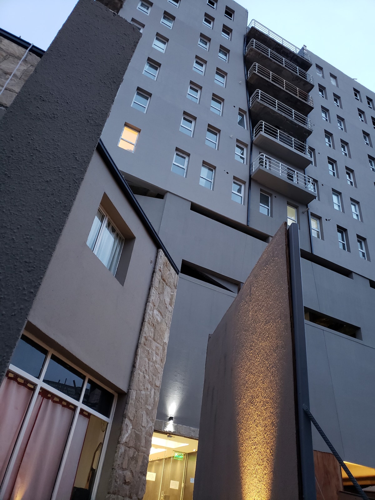 The exterior of a modern multi-story building is depicted, showcasing a combination of gray surfaces and stone accents. A softly illuminated entrance is visible, with balconies outlined against the evening sky. Windows on various levels add to the contemporary architectural design.