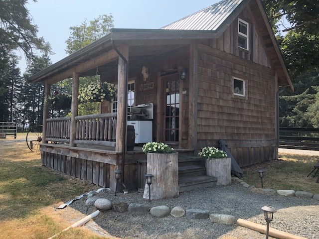 The cabin features a rustic wooden exterior with a welcoming porch adorned with flower pots. A stone pathway leads up to the entrance, highlighting the natural surroundings and spacious outdoor area. Nearby, the lush greenery and horse facilities create a serene atmosphere.