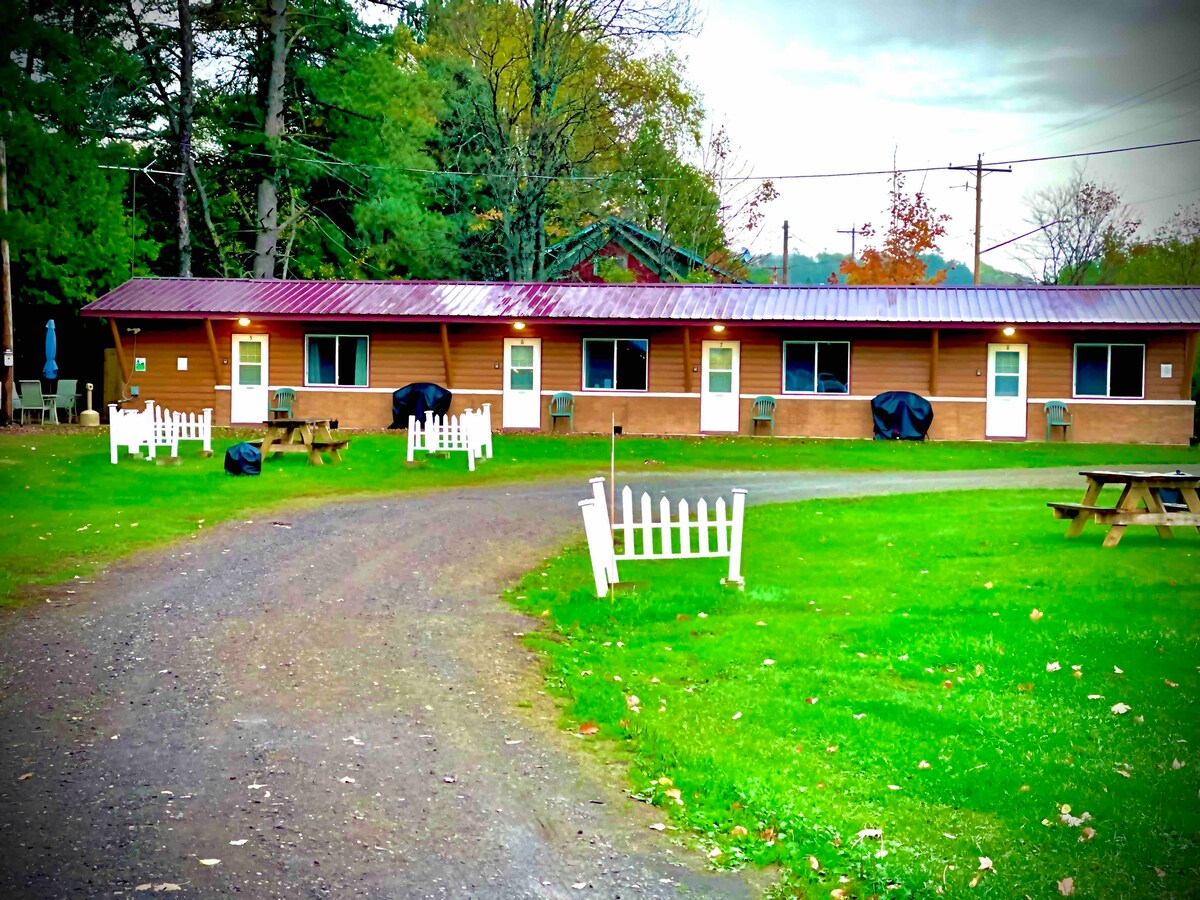 The exterior of a rustic court-style motel is showcased, featuring several doors leading to individual units. A grassy area is visible in front, with a dirt path and picnic tables for outdoor relaxation. Trees surround the property, providing a natural backdrop.