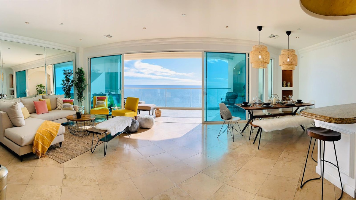 The spacious living area features large glass doors offering panoramic ocean views. A modern sectional sofa and brightly colored armchairs add comfort, while a stylish dining table with seating accommodates guests. A light-colored rug enhances the neutral stone flooring, illuminated by natural light.