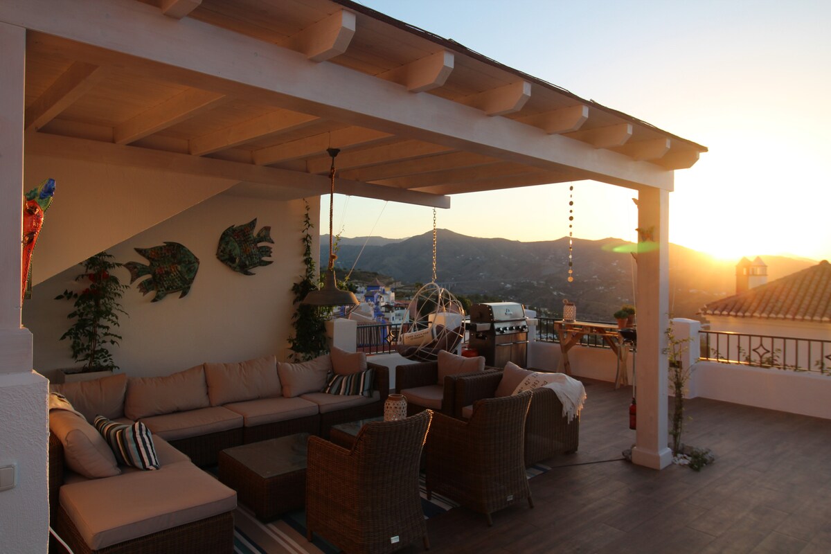 A spacious outdoor seating area features a comfortable lounge set under a wooden pergola. The sun sets in the background, casting a warm glow over the area. Decorative fish hang on the wall, and mountain views are visible beyond the seating.