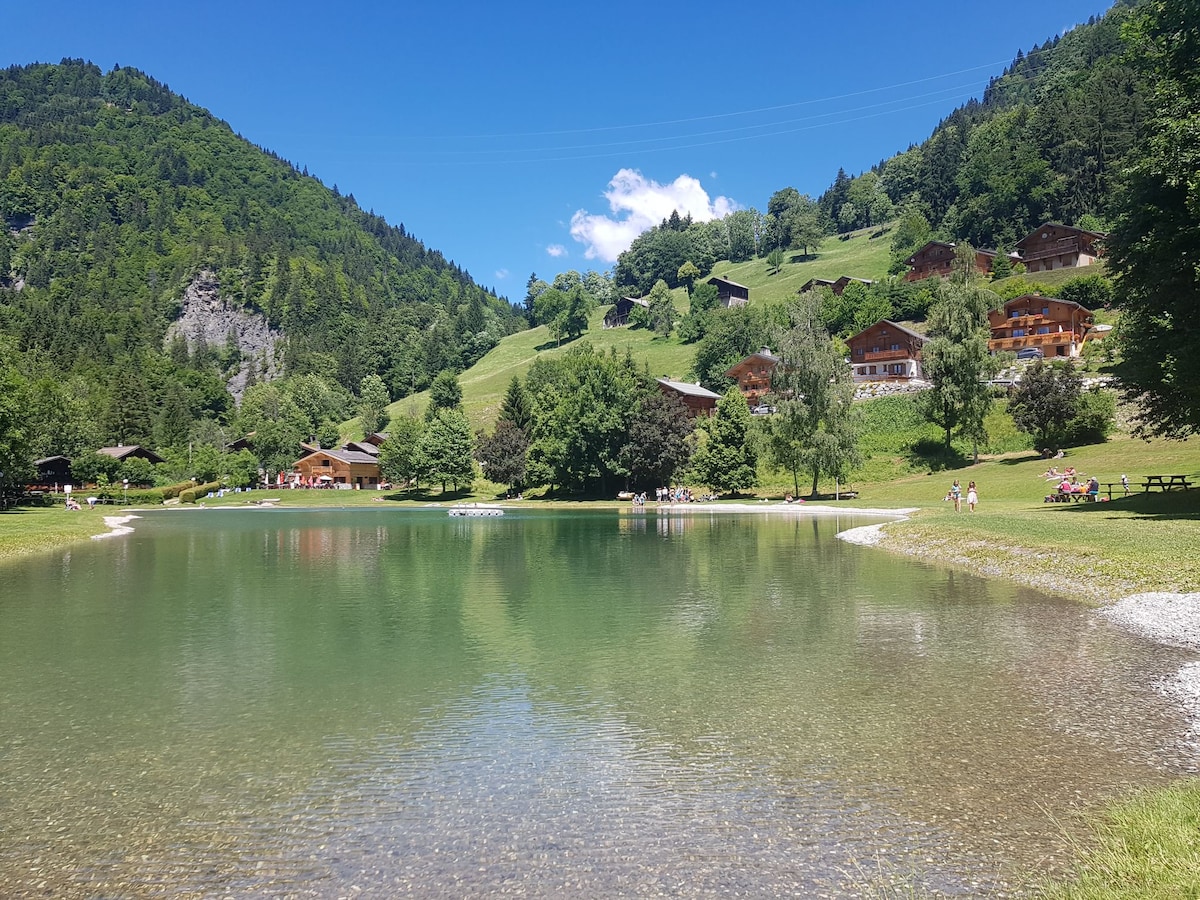A serene lake is captured, reflecting the surrounding lush greenery and blue sky. Wooden chalets are visible on the hillside, and people are seen enjoying the tranquil atmosphere along the water's edge. A few trees offer shade near the lakeside.