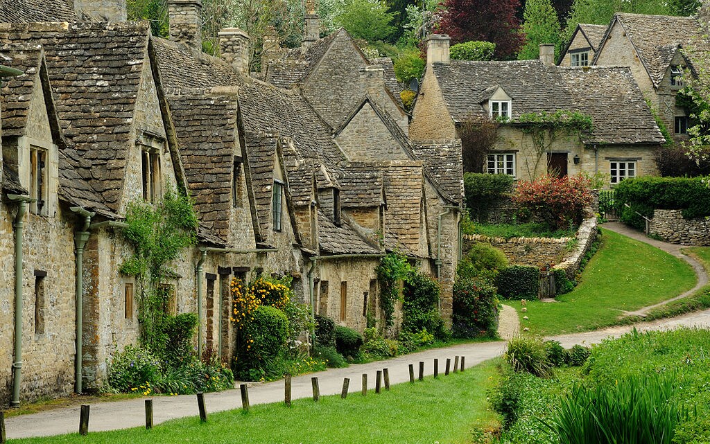 A charming row of stone cottages features traditional architecture characterized by sloping roofs and small windows. Lush greenery surrounds the pathway, which leads past colorful flowerbeds, creating a picturesque village atmosphere. Well-maintained gardens enhance the inviting landscape.