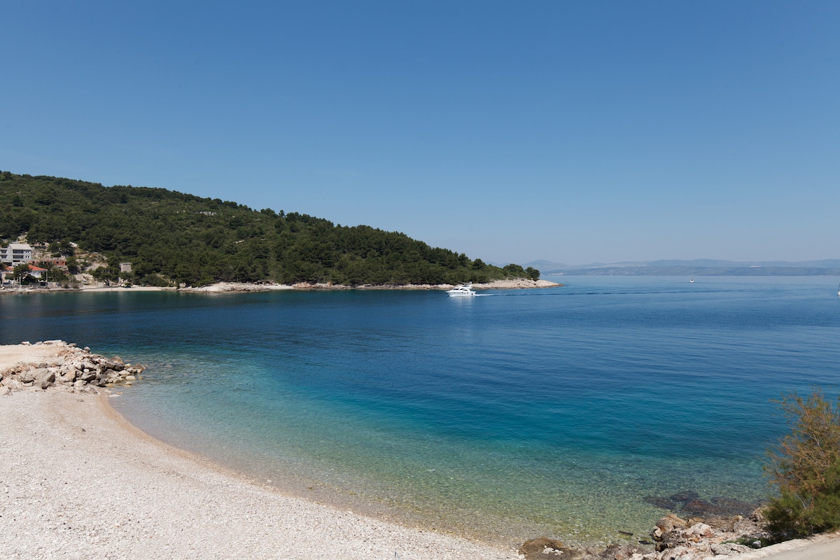 A serene beach scene is presented, featuring clear blue water gently lapping against the pebbled shore. Lush green hills are visible in the background, providing a natural frame to the tranquil bay. The sky is clear with bright sunlight reflecting on the water's surface.