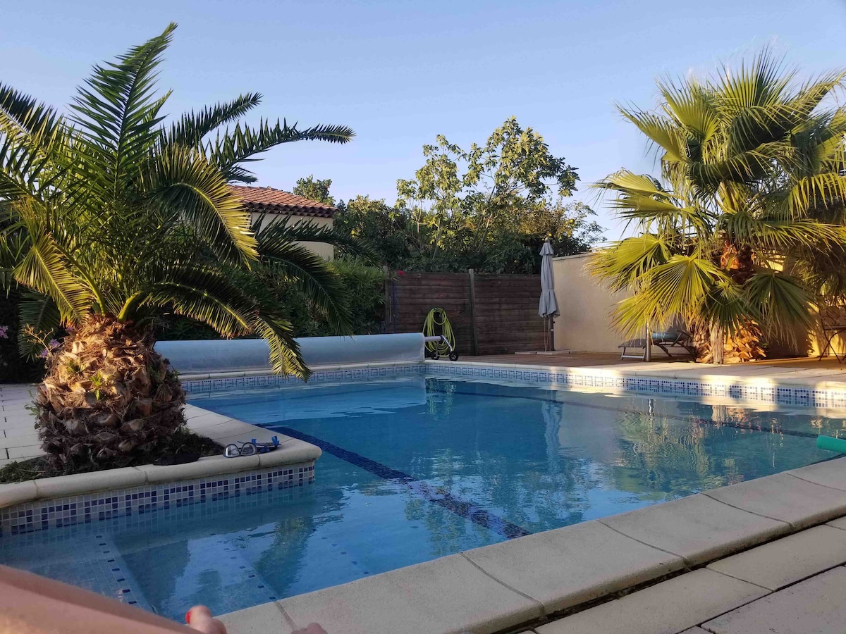 A private swimming pool is surrounded by tropical palm trees and lush greenery. A shaded lounging area is visible in the background, alongside a sunbed. The area is well-maintained, with clear water inviting relaxation under the sun.