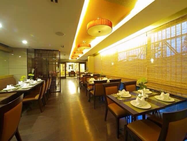 A dining area is presented with wooden tables and brown chairs, evenly spaced throughout the room. A warm glow is emitted from various ceiling lights, complemented by natural light filtering through sheer blinds. Small centerpieces adorn the tables, enhancing the inviting ambiance.