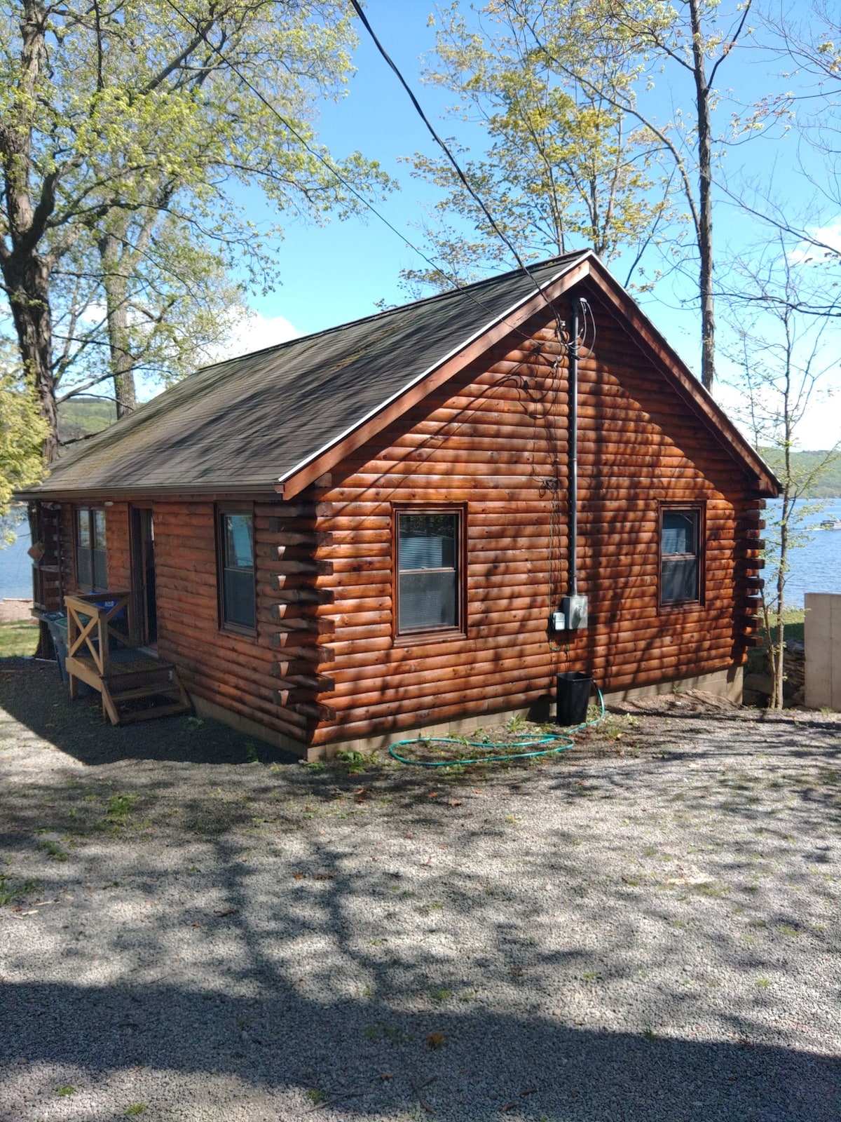 A rustic log cabin sits by the lake, showcasing a warm wooden exterior. Tall trees provide shade, with a gravel pathway leading to the entrance. The tranquil water reflects the natural surroundings, offering a serene backdrop to the cabin.