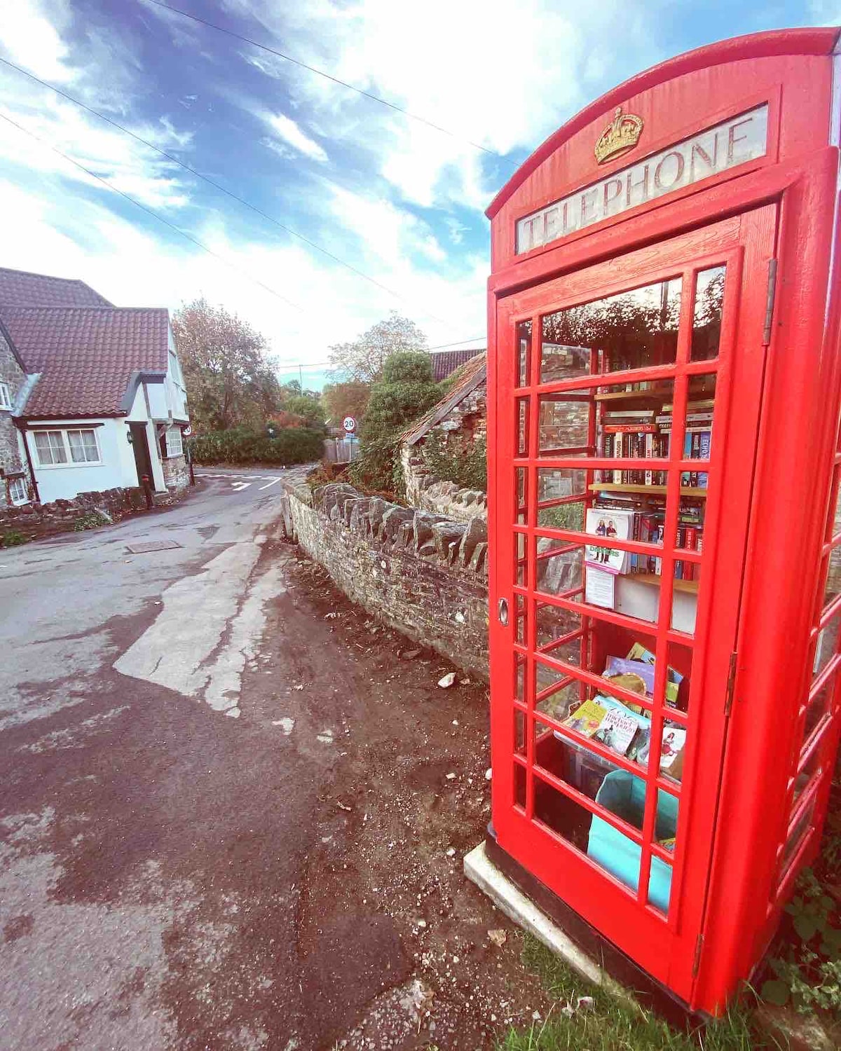 A classic red telephone box stands alongside a narrow road, framed by quaint stone walls and residential buildings. Lush greenery peeks from behind, and the sky is scattered with clouds, adding a touch of serenity to the village scene.