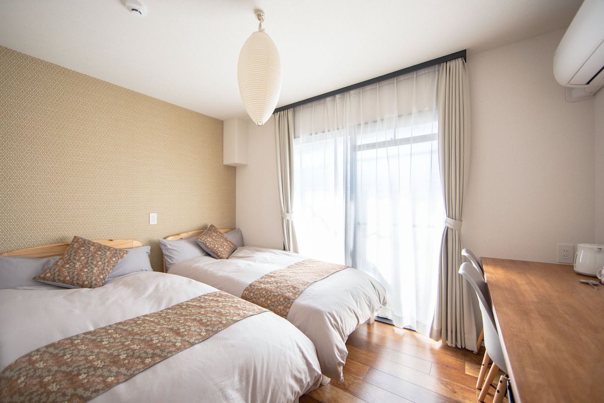 A bright guest room features two spacious beds dressed in light bedding, accented by decorative cushions. Natural light fills the space through a large window, which is framed by sheer curtains. A wooden table with chairs offers a cozy area for work or relaxation.