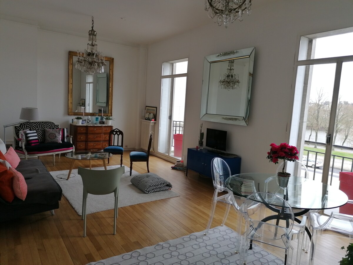 A bright living area is highlighted by large windows offering river views. Elegant chandeliers hang from the ceiling, complementing the mirrored wall. A mix of seating options, including a sofa and dining chairs, surrounds a glass dining table situated on a light wooden floor.