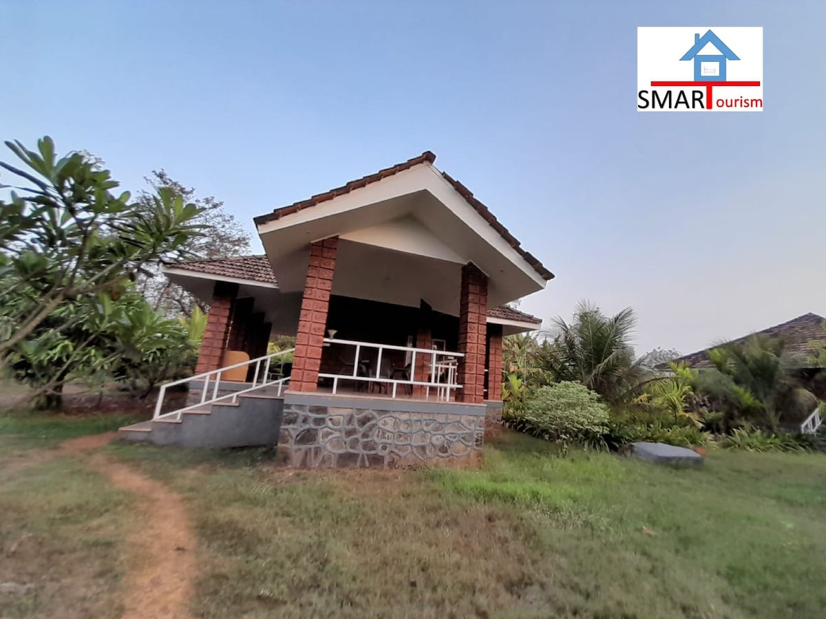 The exterior of a charming house is presented, featuring a combination of stone and brick materials. The structure is surrounded by lush greenery, emphasizing a serene atmosphere. A path leads to the entrance, while a sloped roof adds character to the design.