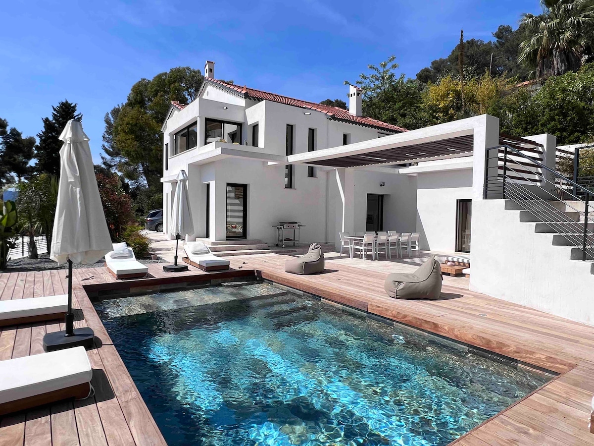 A modern villa with a clean aesthetic is showcased, featuring a heated pool surrounded by wooden decking. Lounge chairs are positioned for relaxation, and a shaded dining area is seen under a pergola. The exterior is bright and inviting against a clear blue sky.